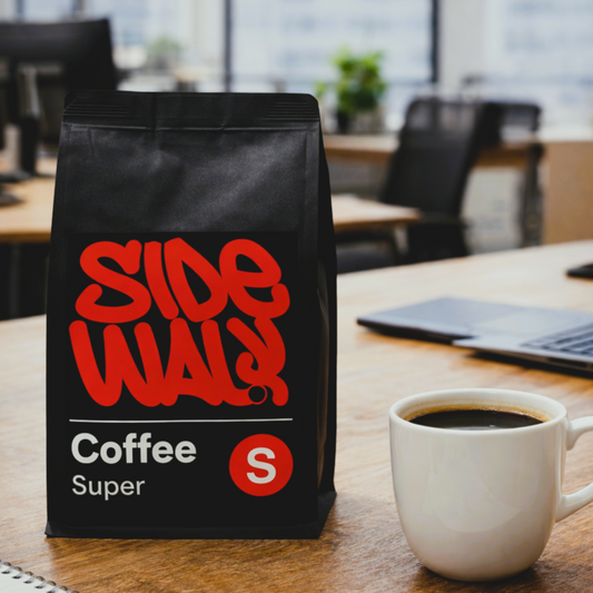 Black coffee bag with red text on a table next to a white mug of coffee, in an office setting.